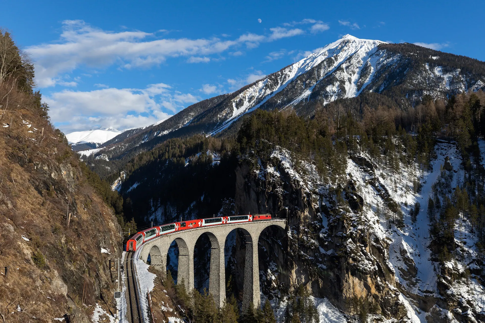 A red Rhaetian Railway train crossing a stone viaduct in the Swiss Alps